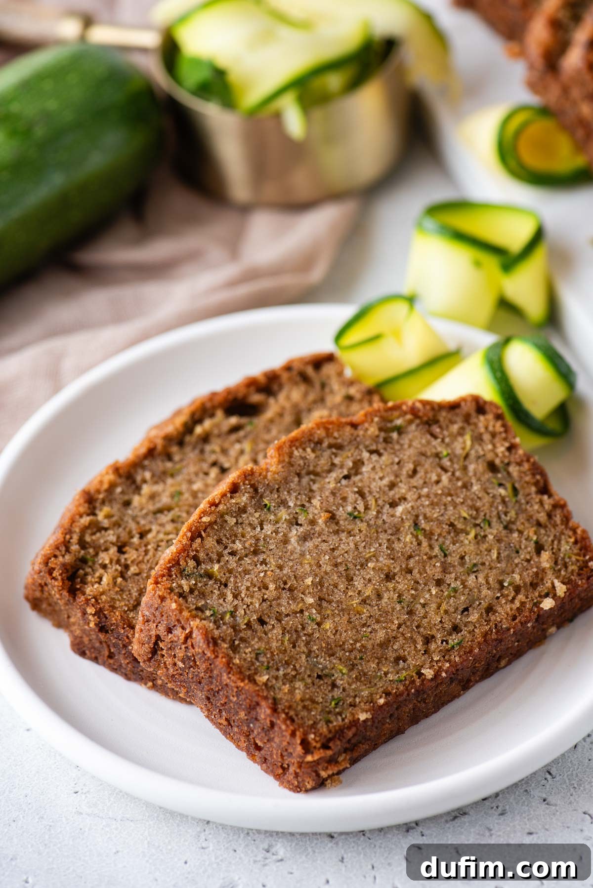 Irresistibly Moist Zucchini Bread 3 Two slices of homemade zucchini bread artfully arranged on a pristine white plate, complemented by fresh slices of zucchini and whole zucchinis, emphasizing its fresh ingredients.