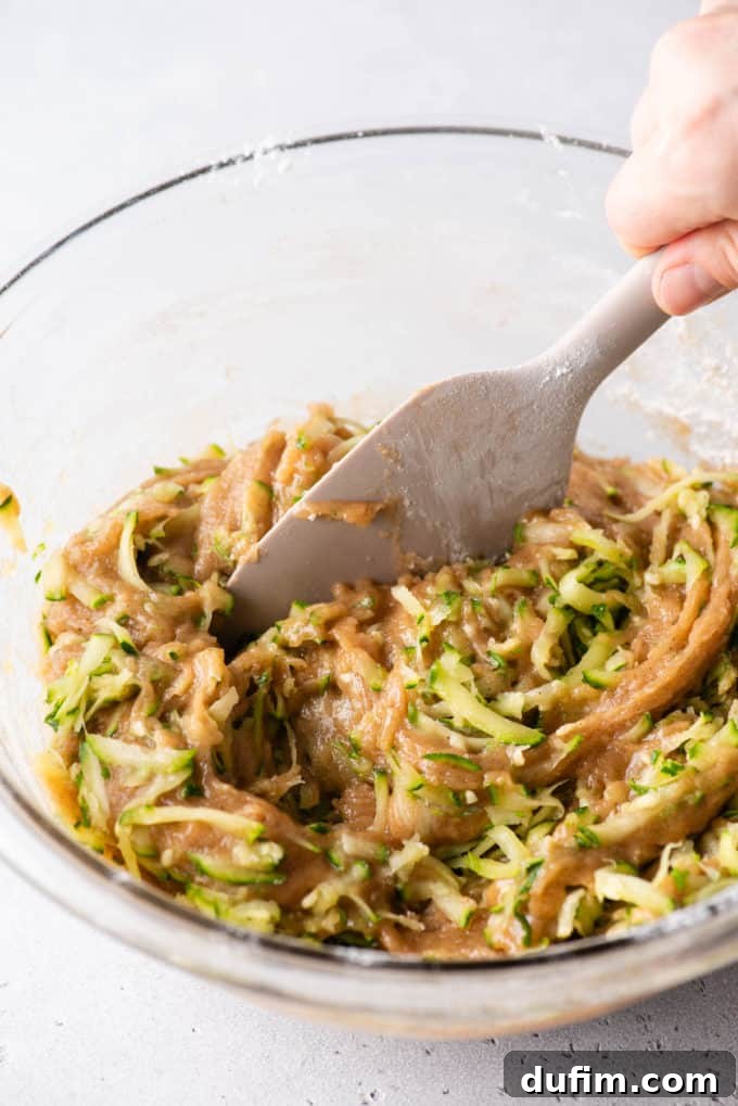 Irresistibly Moist Zucchini Bread 8 Zucchini bread batter being carefully mixed in a clear glass bowl with a tan spatula, showing the rich texture before baking.