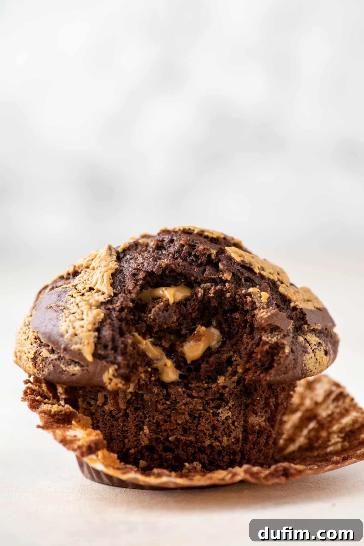 A single, perfectly baked chocolate peanut butter muffin, with its paper liner partially peeled, showcasing its irresistibly moist interior and impressive, tall dome.
