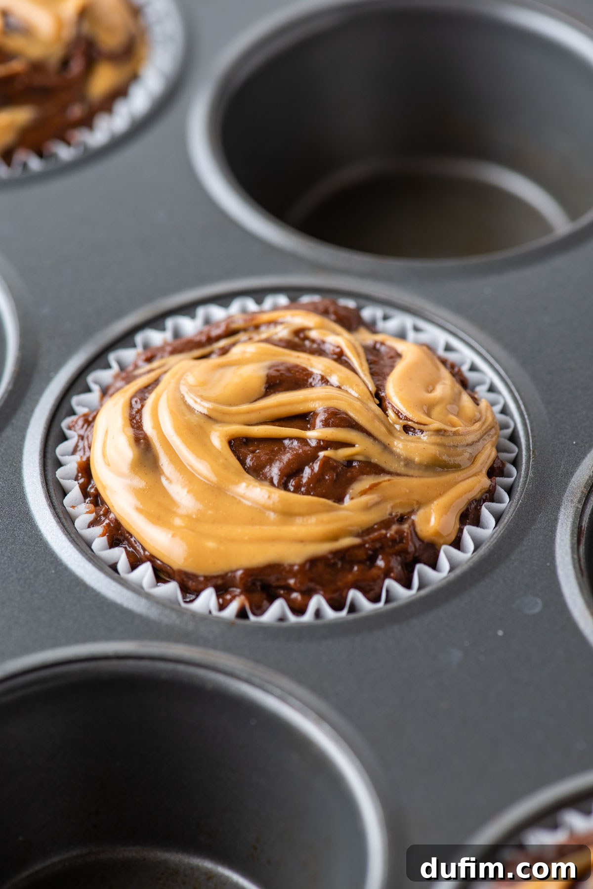 Close-up of unbaked chocolate peanut butter muffin batter with visible peanut butter swirls, sitting in a muffin tin, patiently waiting to be placed in the oven.