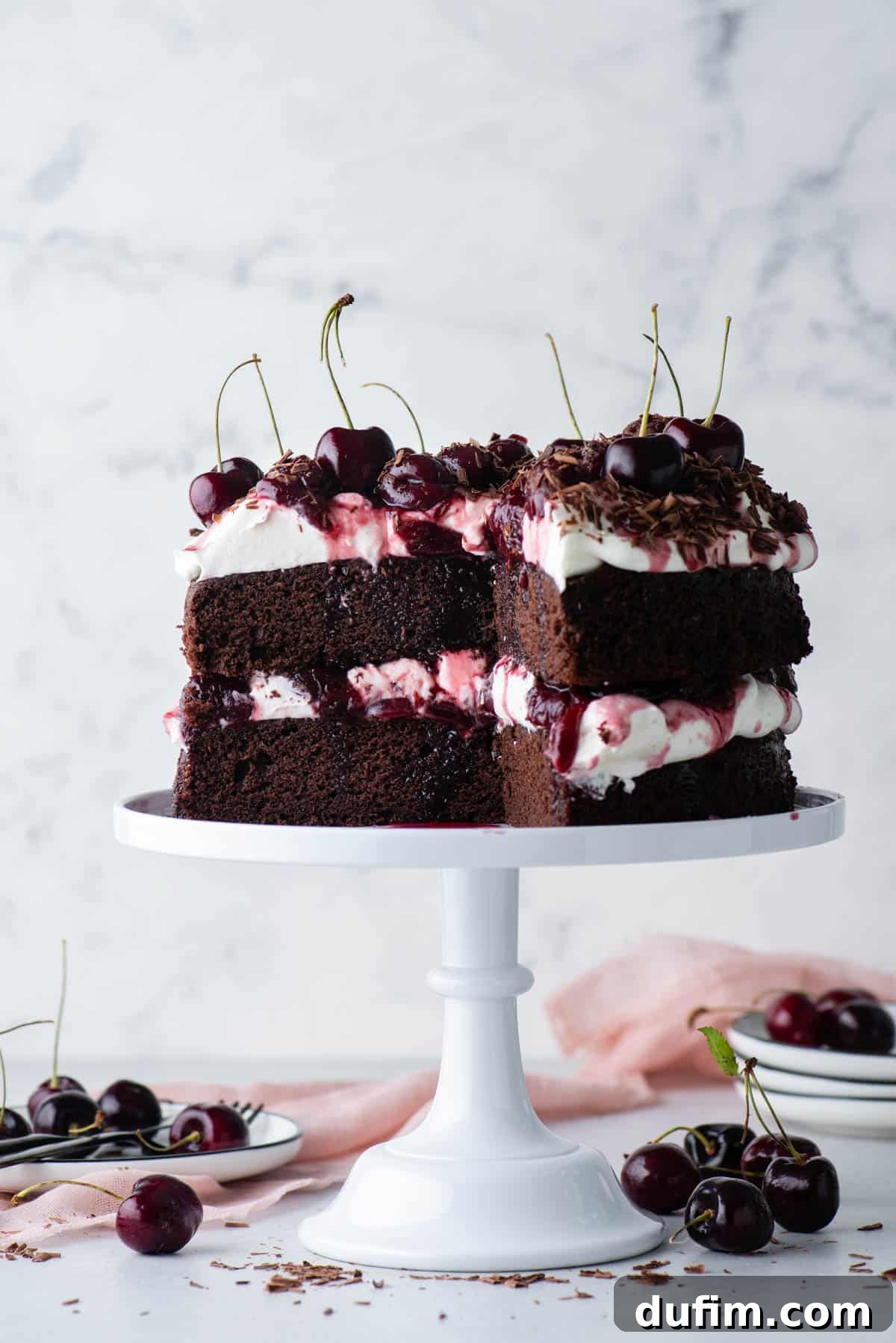 Decadent Black Forest Cherry Torte 2 black forest cake on a white cake stand with a large slice taken out, with stacks of white plates and cherries scattered around