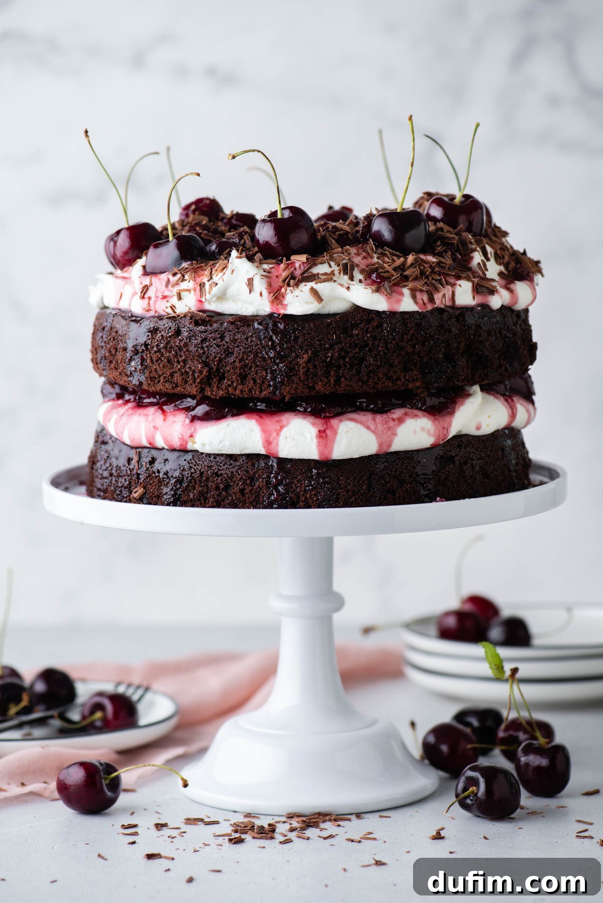 Decadent Black Forest Cherry Torte 13 black forest cake on a white cake stand with stacks of white plates and cherries surrounding it