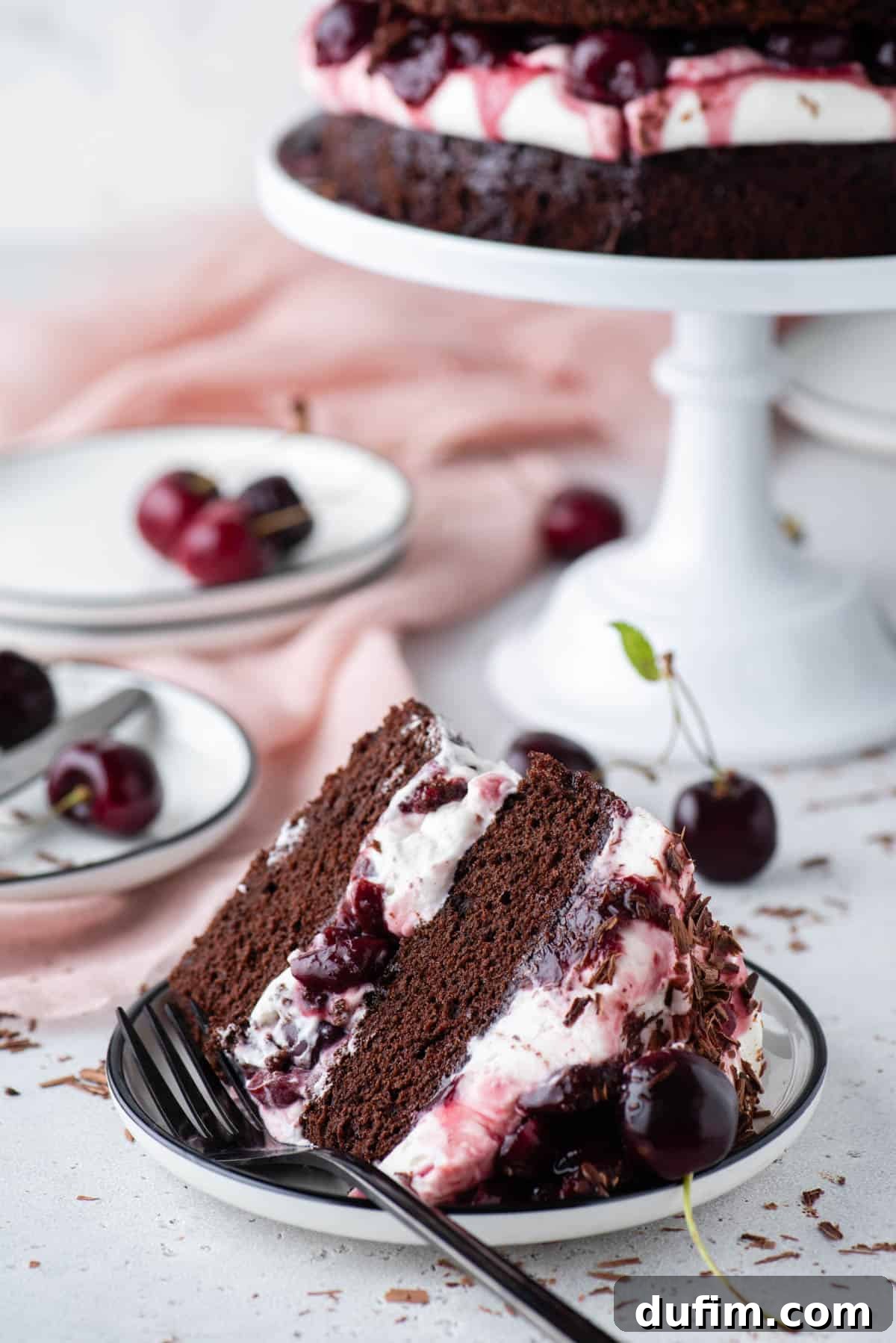 Decadent Black Forest Cherry Torte 4 a slice of black forest cake on a white plate with a form with a cake stand and more plates in the background and cherries and chocolate shavings scattered around