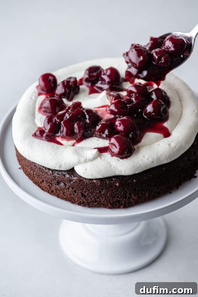 Decadent Black Forest Cherry Torte 8 one layer of chocolate cake on a white cake stand with stabilized whipped cream on top and a cherry filling being spooned on top of it