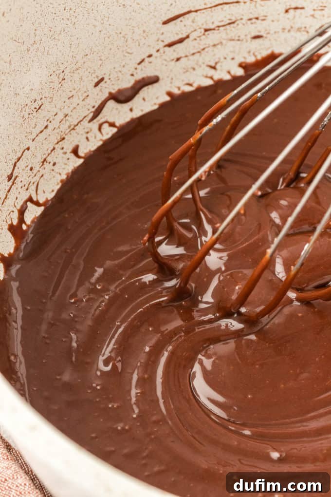 A close-up view of the chocolate frosting being vigorously whisked in a saucepan, ensuring all powdered sugar clumps are smoothed out.