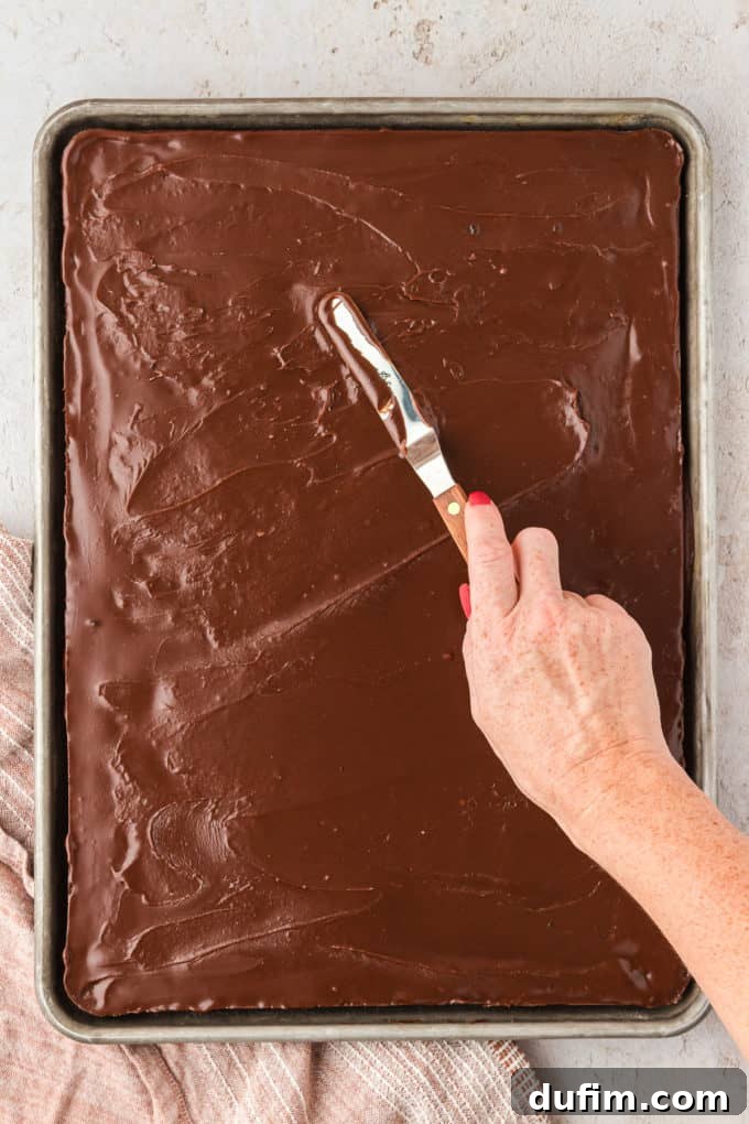 Chocolate frosting being expertly spread across the warm Texas Sheet Cake with a butter knife, ensuring even coverage.