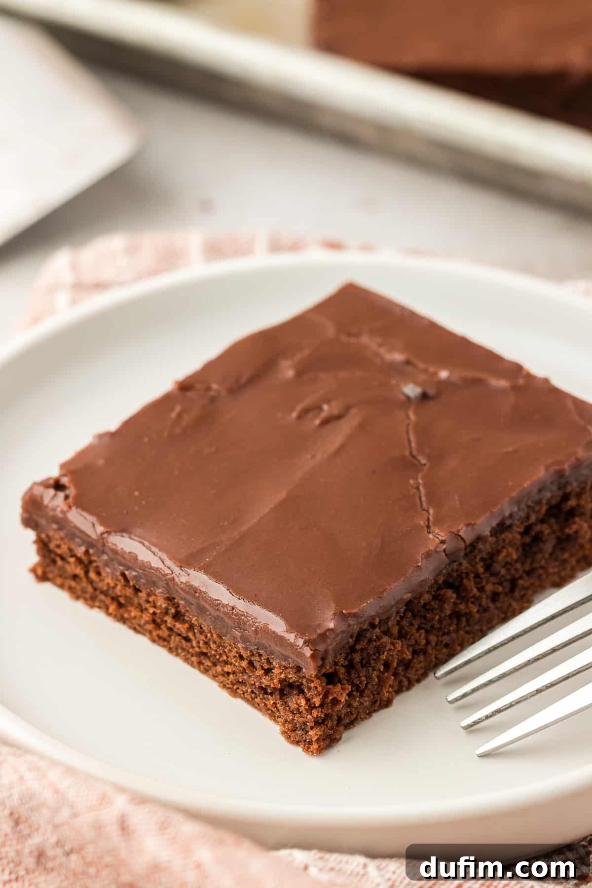 A single slice of Texas Sheet Cake on a white plate with a fork, ready to be enjoyed, accompanied by a neatly folded tan kitchen towel.