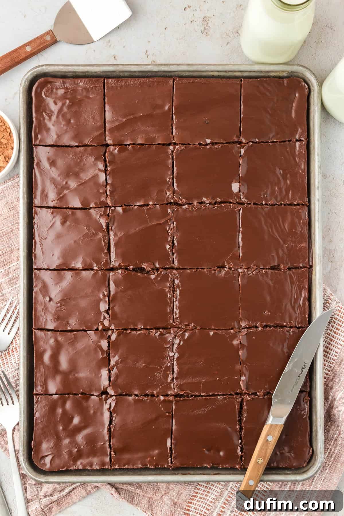 A large sheet pan of Texas Sheet Cake cut into neat squares, with a knife resting on top, surrounded by a spatula, a bowl of cocoa powder, milk, and a fork, against a tan kitchen towel.