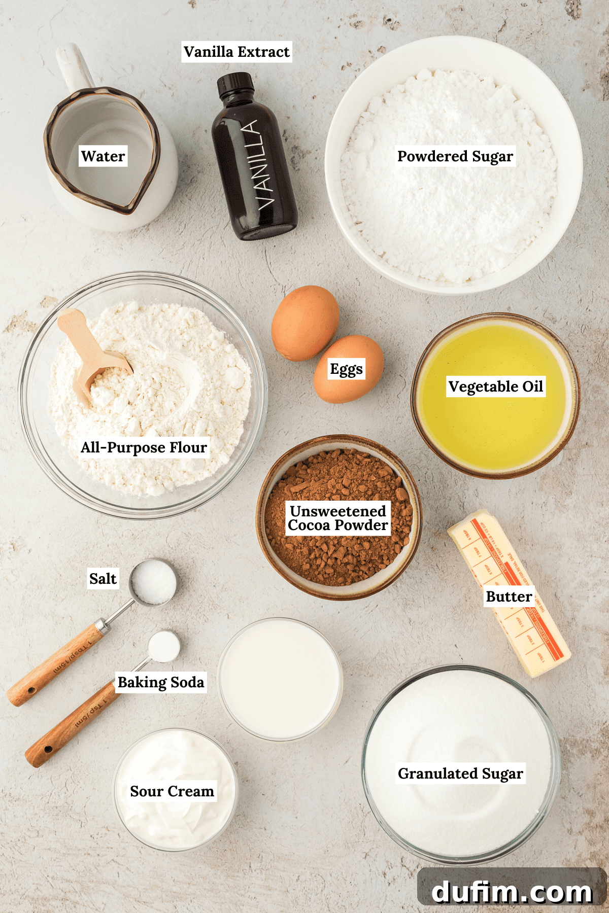 All the essential ingredients for Texas Sheet Cake laid out: water, vanilla extract, powdered sugar, all-purpose flour, vegetable oil, unsweetened cocoa powder, butter, salt, baking soda, sour cream, granulated sugar, and milk.