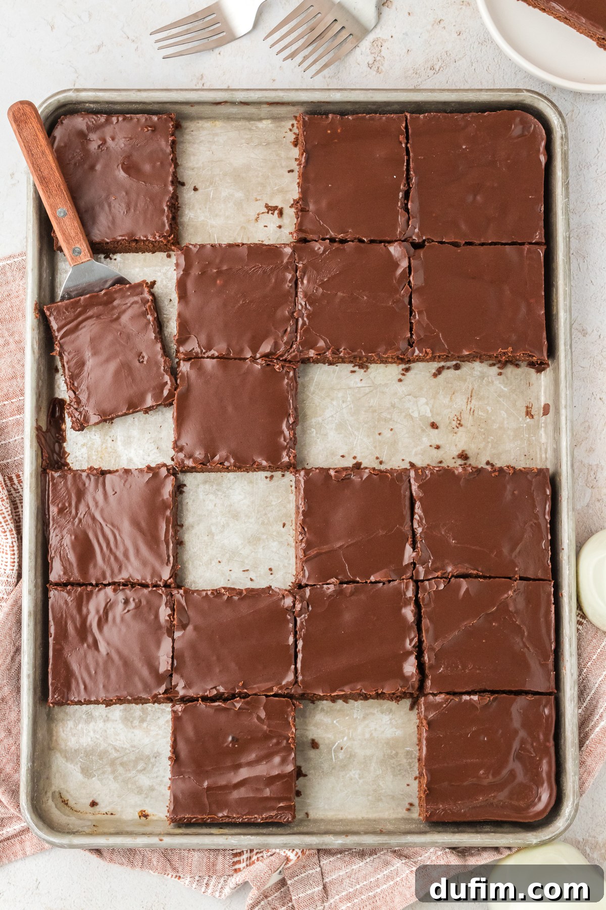 Square slices of Texas Sheet Cake beautifully arranged on a large sheet pan, with a few pieces already taken, and a spatula poised to serve another slice. A plate with a slice and forks complete the scene.