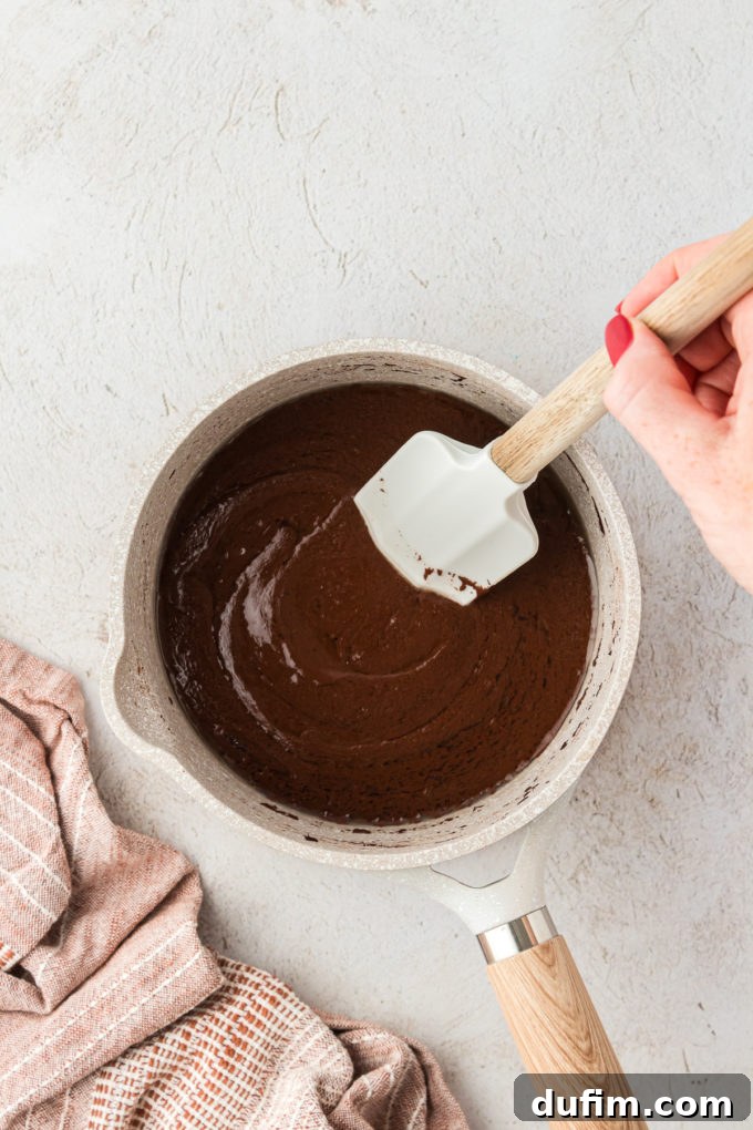 The wet ingredients for Texas Sheet Cake (water, oil, cocoa) being mixed in a saucepan with a red spatula, ensuring the cocoa dissolves evenly.
