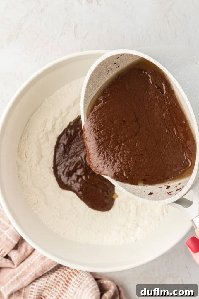 The warm chocolate mixture being poured from a saucepan into a white bowl containing the dry flour mixture for the Texas Sheet Cake.