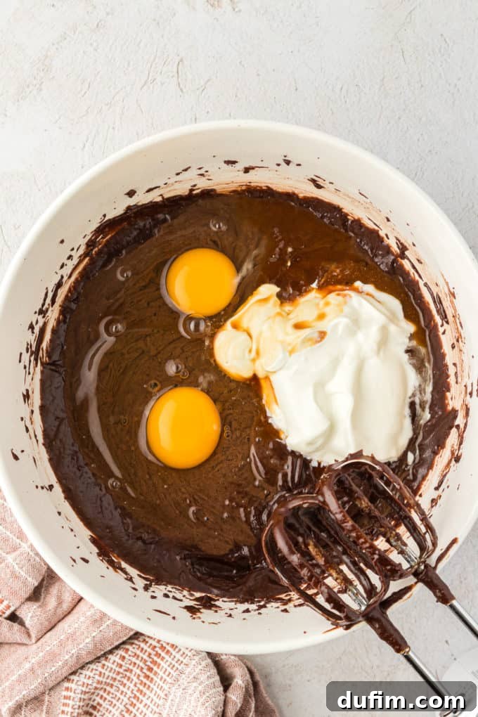 Raw eggs and a dollop of sour cream added to the Texas Sheet Cake batter in a white bowl, with mixer beaters covered in chocolate batter leaning on the bowl.