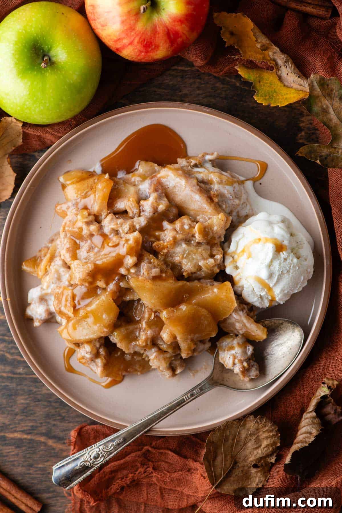 A slice of golden-brown apple crisp on a white plate, generously drizzled with caramel sauce, accompanied by a scoop of melting vanilla ice cream and a spoon. Fresh apples, vibrant fall leaves, and an orange linen towel create a rustic autumnal backdrop.