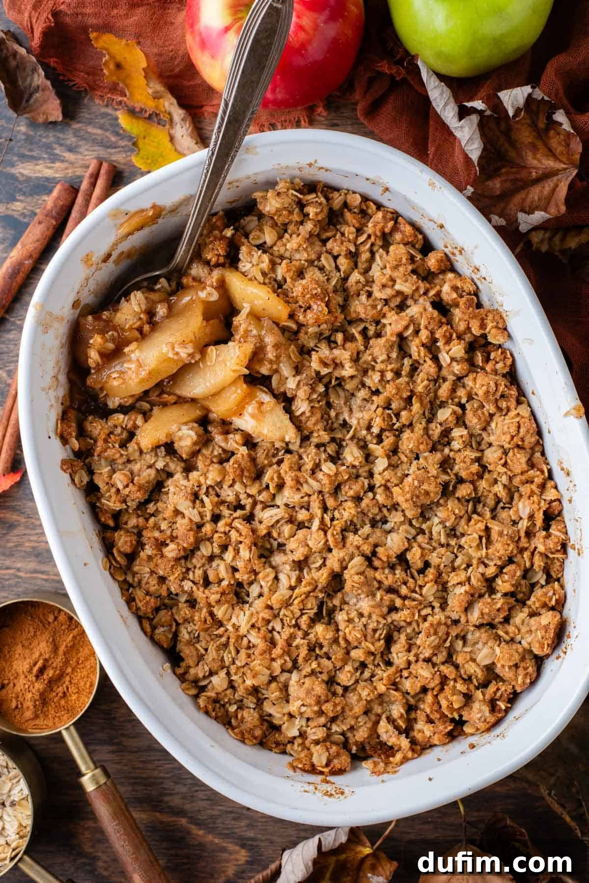 A white baking dish filled with freshly baked, steaming apple crisp, with a spoon firmly planted within its golden-brown topping. The dish is beautifully framed by warm fall leaves, aromatic cinnamon sticks, and a deep orange linen towel, evoking the cozy essence of autumn.