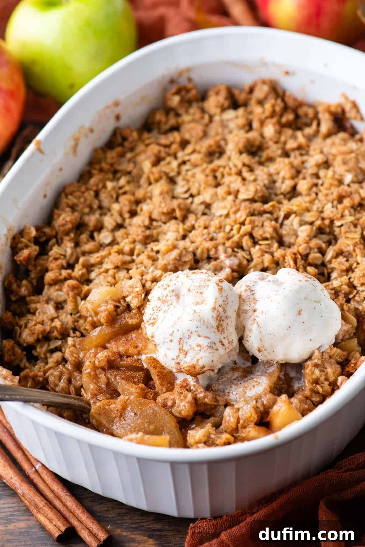 A white baking dish, fresh from the oven, showcasing a warm apple crisp with a spoon and two melting scoops of creamy vanilla ice cream resting on top. The steam gently rises, highlighting the warmth and deliciousness of the dessert.