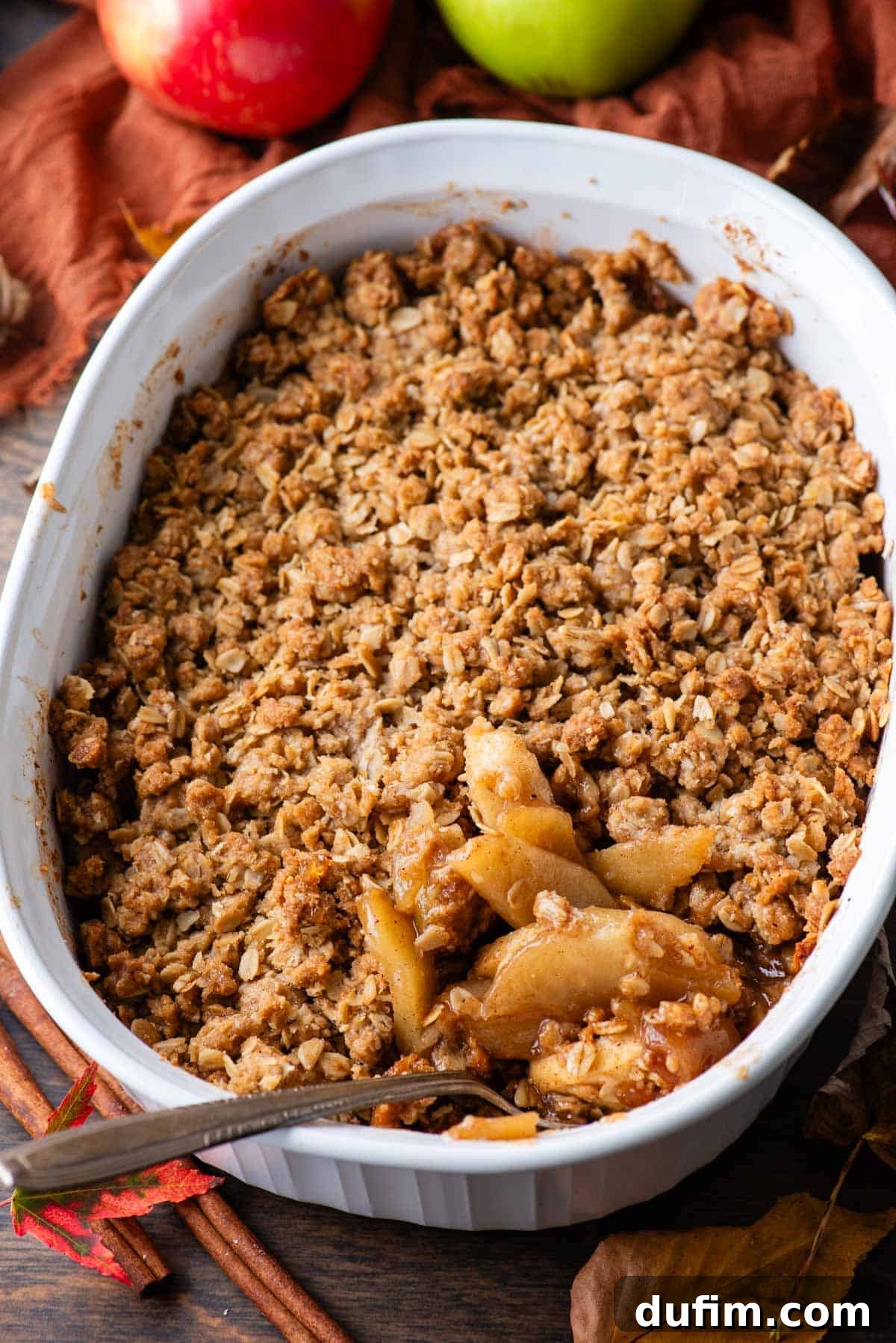 A white baking dish full of perfectly baked apple crisp, with a serving spoon nestled within. The inviting scene is completed by cinnamon sticks, a dark orange linen towel, one whole red apple, one whole green apple, and a scattering of vibrant fall leaves, highlighting the seasonal delight.