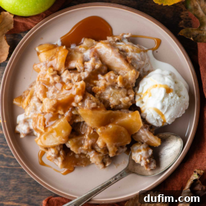 A slice of golden-brown apple crisp on a white plate, generously drizzled with caramel sauce, accompanied by a scoop of melting vanilla ice cream and a spoon. Fresh apples, vibrant fall leaves, and an orange linen towel create a rustic autumnal backdrop.
