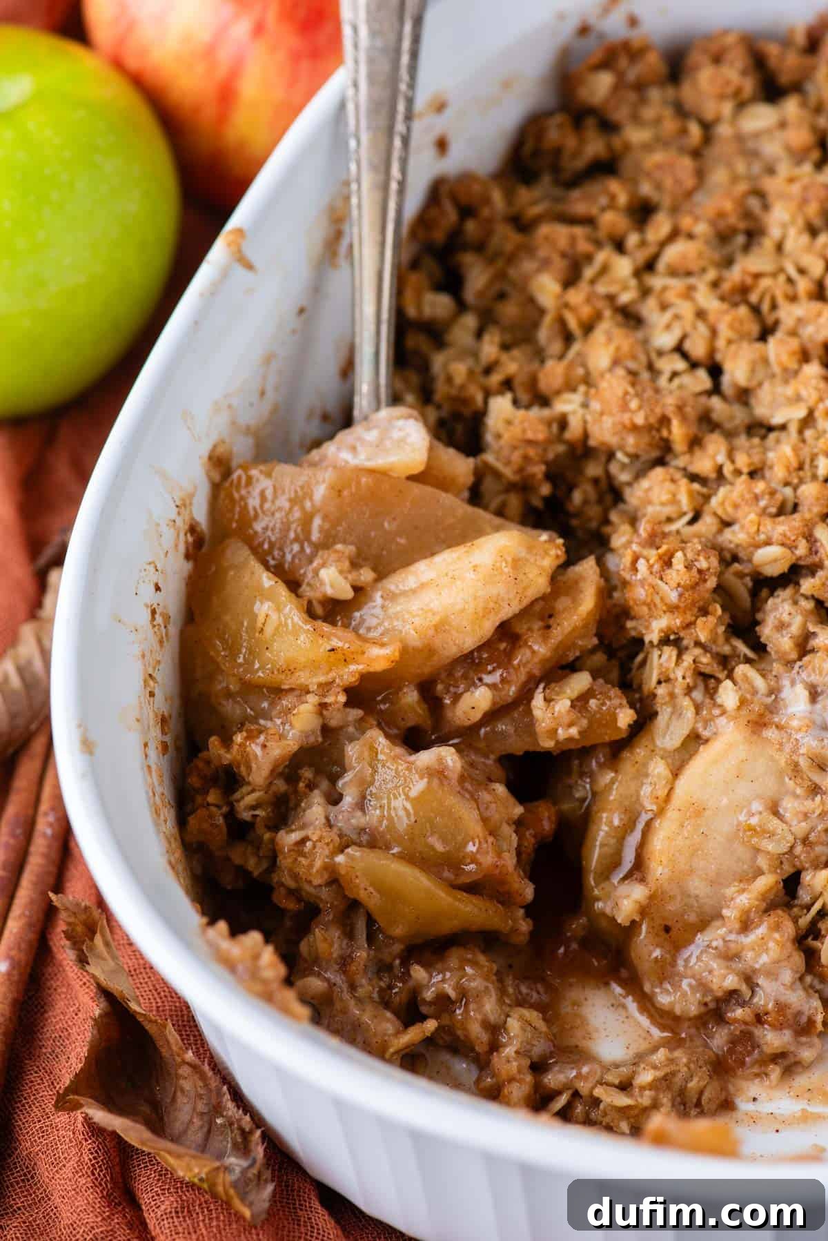 A close-up view of a white rectangular baking dish filled with a golden-brown apple crisp, a spoonful missing, revealing the tender apple filling beneath. A dark orange kitchen towel and a whole green apple rest beside the dish, emphasizing the freshness and warmth of the dessert.