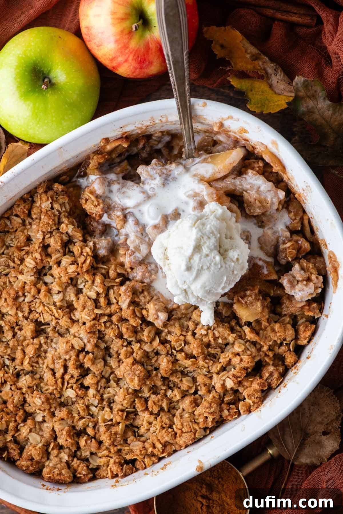 A white baking dish brimming with freshly baked apple crisp, featuring a spoon and a melting scoop of vanilla ice cream nestled within. A vibrant green apple, autumn leaves, and a dark orange linen towel complete the inviting scene, highlighting the seasonal appeal of the dessert.