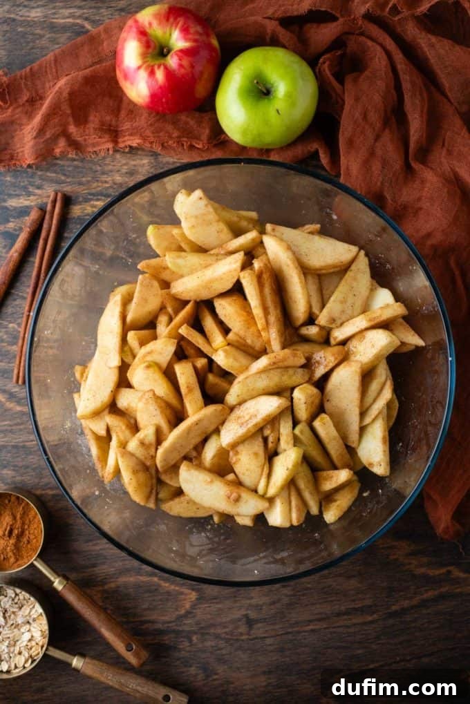 A clear glass bowl filled with neatly sliced apples, surrounded by cinnamon sticks, a whole green apple, a spoonful of ground cinnamon, an orange linen towel, and scattered fall leaves, setting a warm and inviting scene for baking.