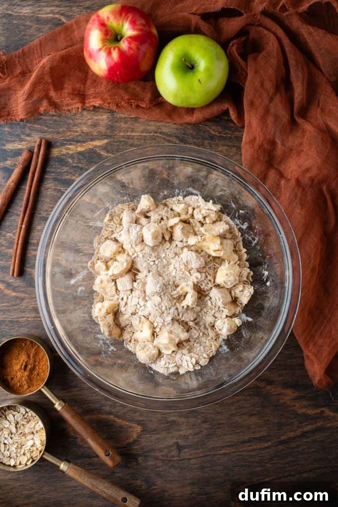 Ingredients for the apple crisp crumble topping, including old-fashioned rolled oats, flour, brown sugar, cinnamon, and salt, artfully arranged in a clear glass bowl on a rustic wooden surface. A spoon of oats, a spoon of cinnamon, cinnamon sticks, a whole green apple, and a dark orange towel complete the cozy baking scene.
