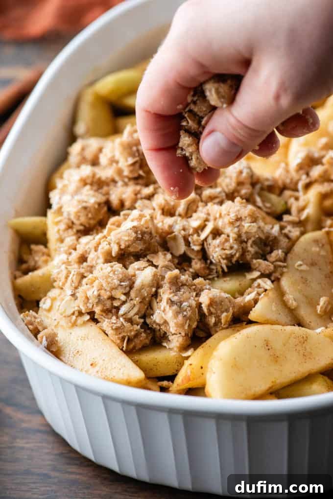 A white baking dish filled with a cinnamon-spiced apple mixture, being generously topped with an oat crumble, ready for baking.