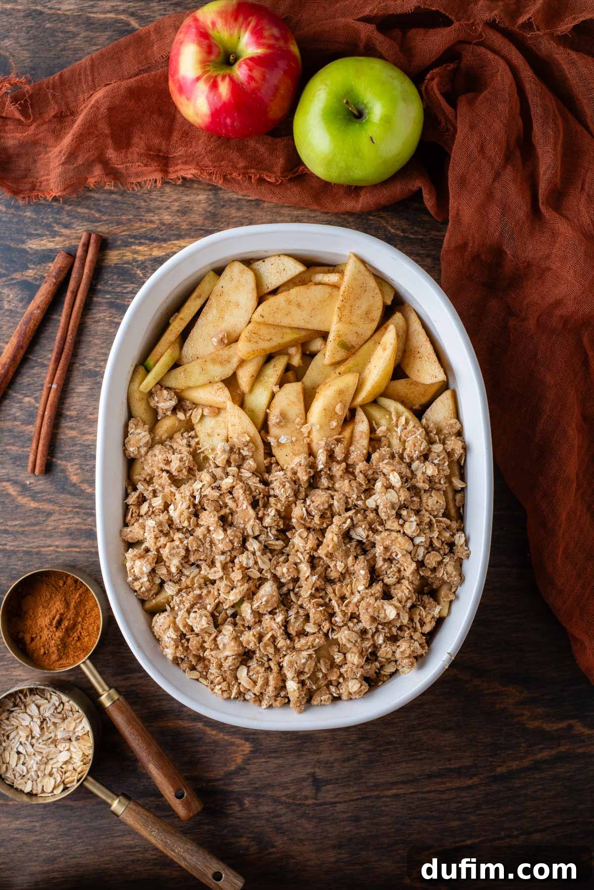 A white baking dish showcases a partially covered apple crisp, with over half of the spiced apple filling already blanketed by the golden oat crumble topping. Surrounding the dish are charming elements like a spoon of oats, a spoon of cinnamon, aromatic cinnamon sticks, a vibrant whole green apple, and a cozy dark orange kitchen towel, illustrating the baking process.