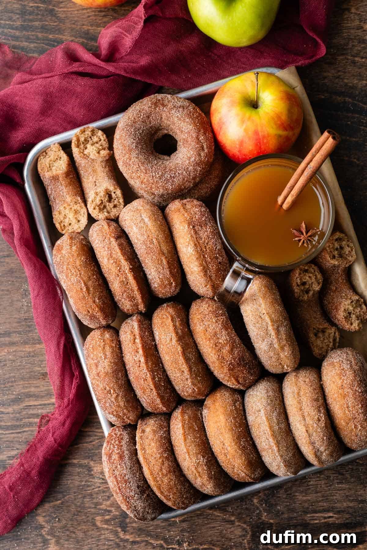 A sheet pan generously filled with golden-brown baked apple cider donuts, some arranged neatly, some stacked, and a few halved. A glass of apple cider with a cinnamon stick and fresh red and green apples rest on a rustic maroon towel, capturing the cozy essence of fall.