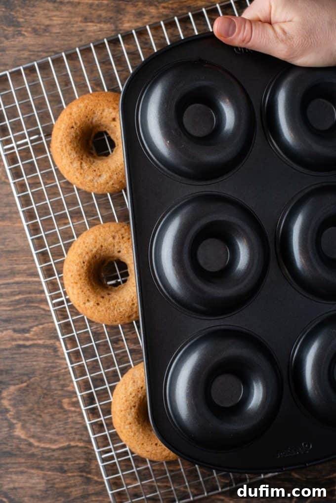 A donut pan being expertly flipped upside down over a wire cooling rack, releasing perfectly baked apple cider donuts onto the rack, indicating they're ready for the next step.