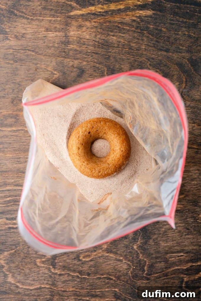 A baked apple cider donut carefully placed inside a ziplock bag, which contains a generous cinnamon sugar mixture, ready for coating on a wooden surface.