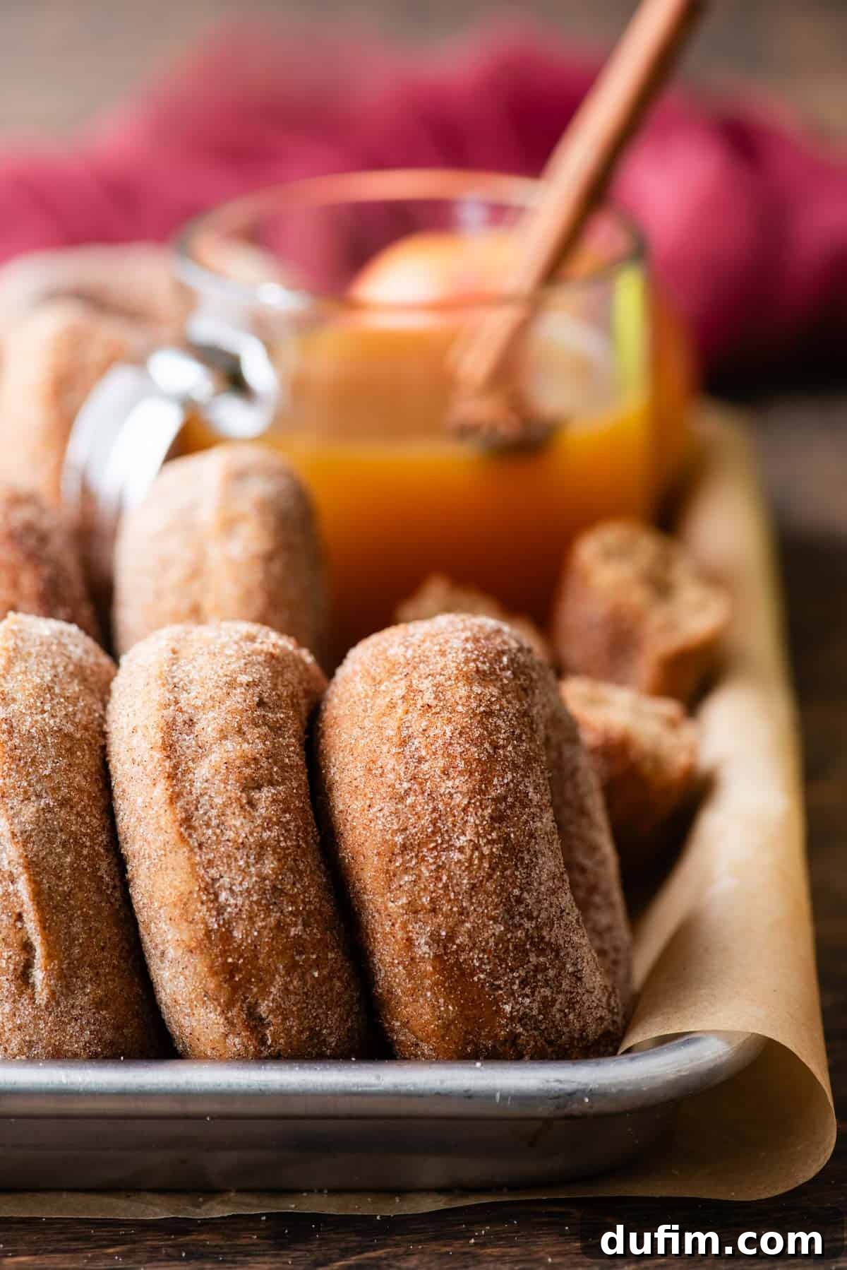 A parchment-lined sheet pan proudly showcases a row of freshly baked apple cider donuts, neatly lined up on their sides. A refreshing cup of apple cider waits patiently in the background, completing a delightful fall scene.