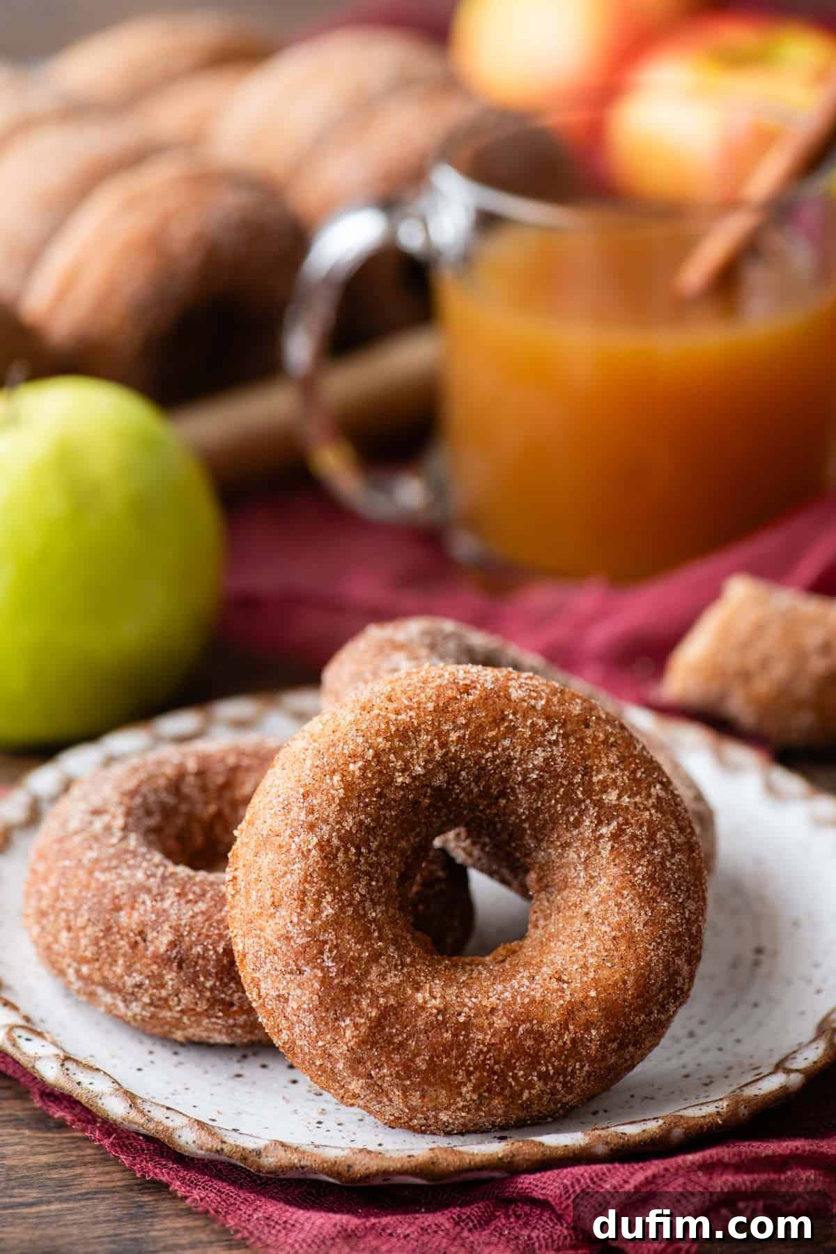 Three golden-brown baked apple cider donuts artfully arranged on a white plate, positioned on a cozy maroon kitchen towel. The scene is completed with a vibrant whole green apple, an inviting cup of apple cider, and additional donuts softly blurred in the background, evoking a comforting autumnal ambiance.