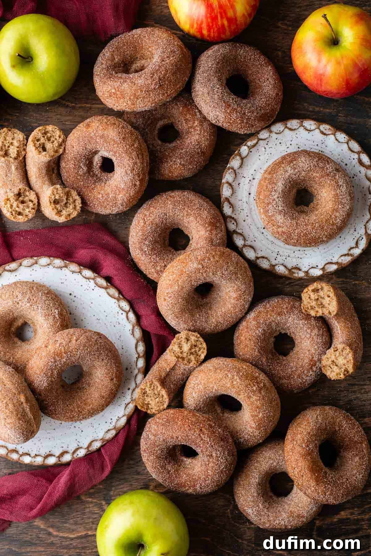 An inviting autumnal scene featuring an array of baked apple cider donuts scattered artfully across a rustic wooden surface. Some are arranged on various white plates, some are thoughtfully halved, and others remain whole. Fresh, vibrant whole apples and a cozy maroon towel complete the rustic tableau, evoking a deep sense of fall comfort and indulgence.