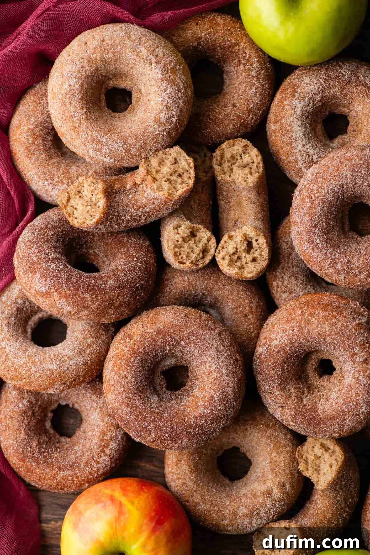 A generous, inviting pile of baked apple cider donuts, including several thoughtfully placed half donuts, artfully arranged on a soft maroon towel. A vibrant whole green apple rests nearby, enhancing the cozy and abundant autumnal theme.