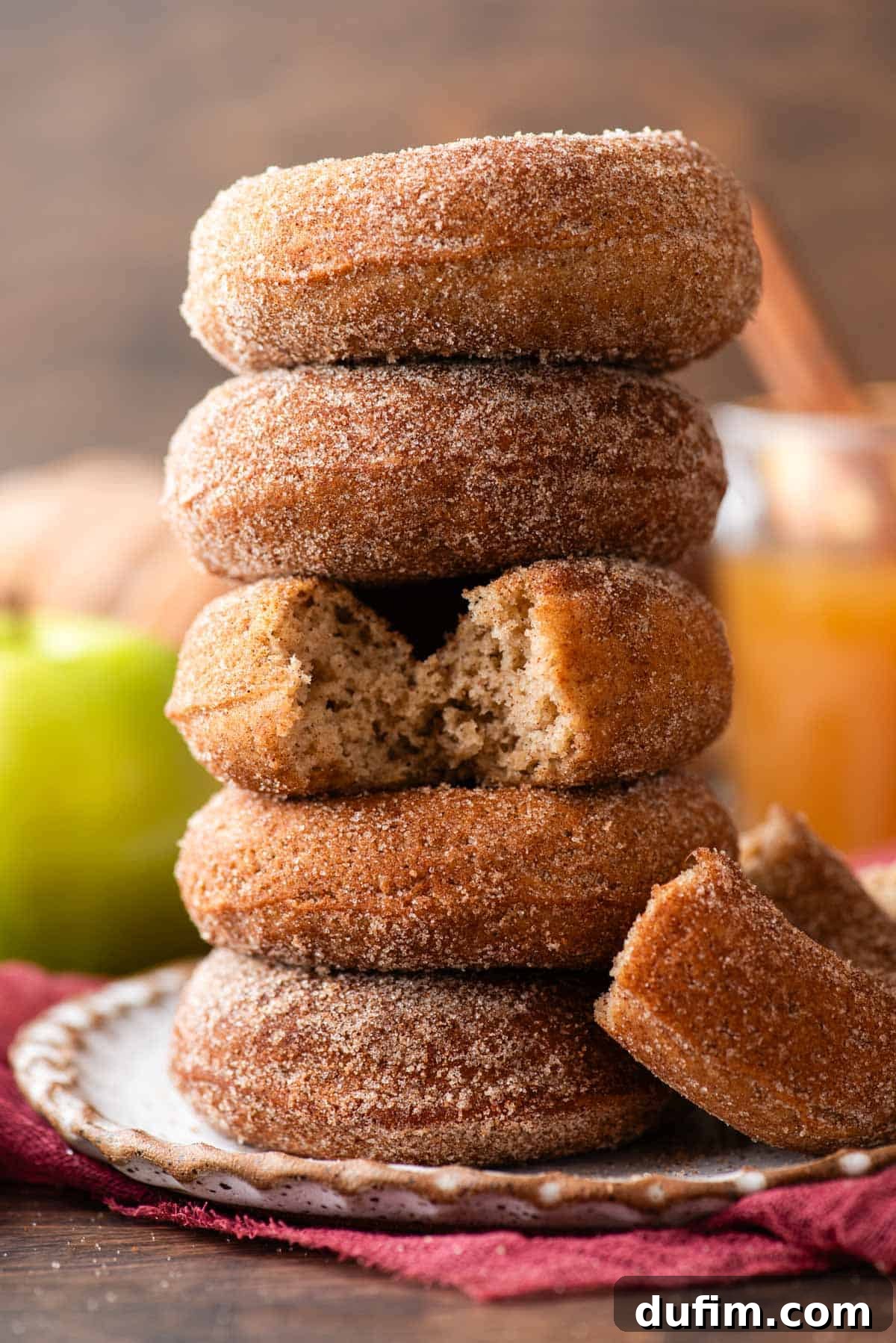 A beautifully stacked trio of baked apple cider donuts on a plate, with a half-eaten donut leaning invitingly beside them, all resting on a cozy maroon towel. A vibrant green apple and a clear glass of apple cider are softly blurred in the background, completing a serene autumnal scene.