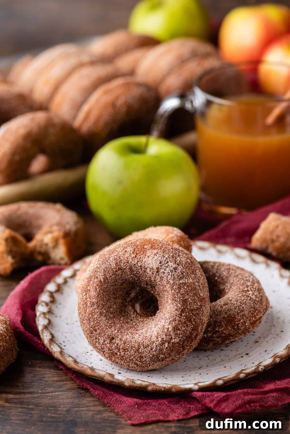 Two perfectly coated baked apple cider donuts nestled on a white plate atop a maroon kitchen towel, surrounded by more freshly baked donuts, a vibrant whole green apple, and a steaming cup of apple cider in the background, creating a warm and inviting autumnal tableau.