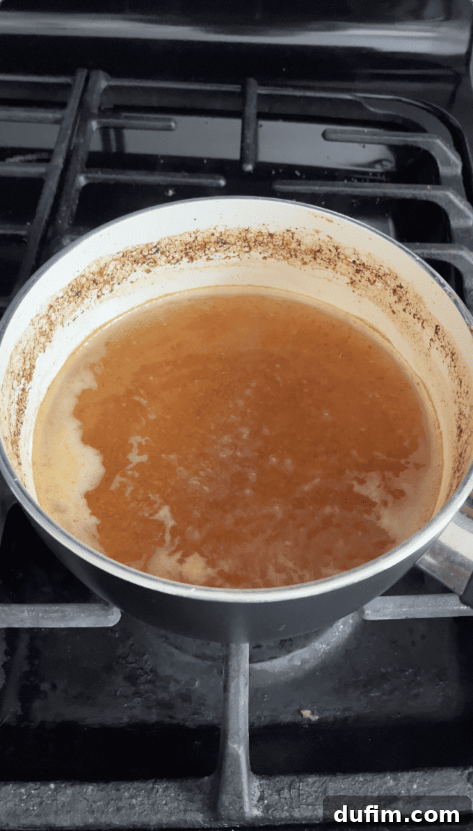 The reduced apple cider, now significantly concentrated and darker, rests in the white saucepan on the oven, poised to be measured and fully cooled for incorporation into the donut recipe.