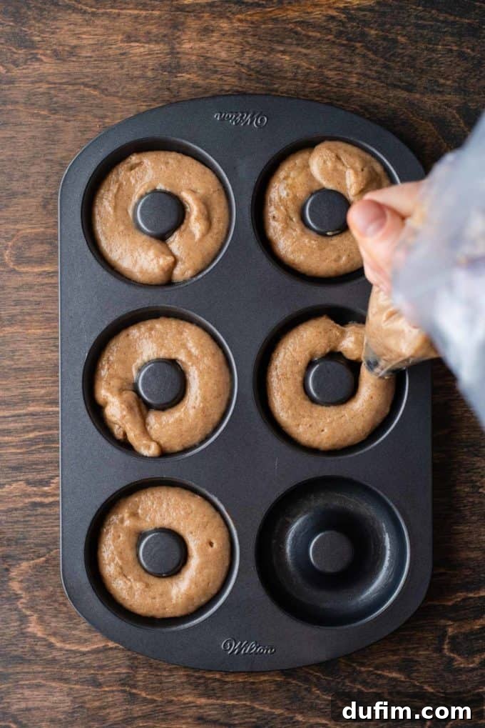 Thick apple cider donut batter being expertly piped from a ziplock bag into the carefully prepared wells of a donut pan, resting on a rustic wooden surface, ensuring even distribution.