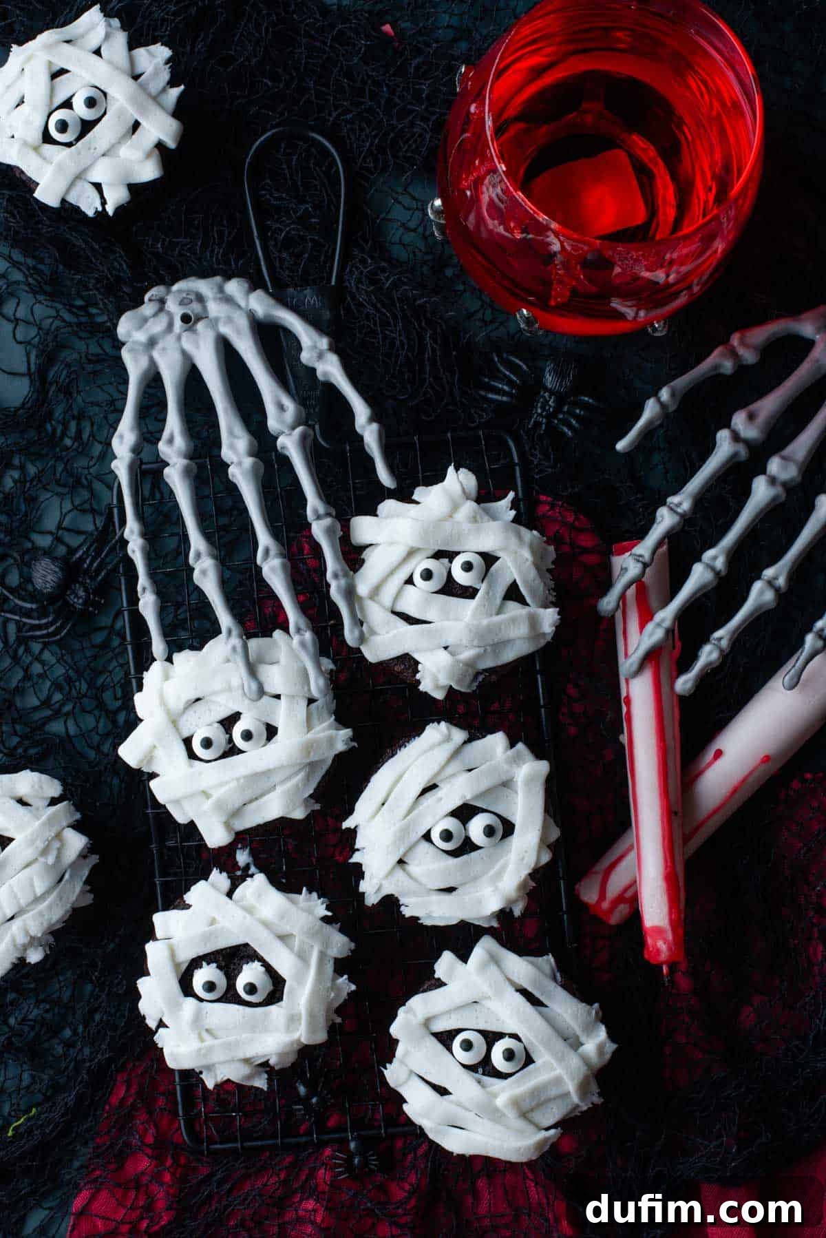 Delicious chocolate mummy cupcakes arranged on a black and red surface, surrounded by Halloween decorations like a glass of red liquid, a skeleton hand, and tiny black plastic spiders. Perfect for spooky celebrations.