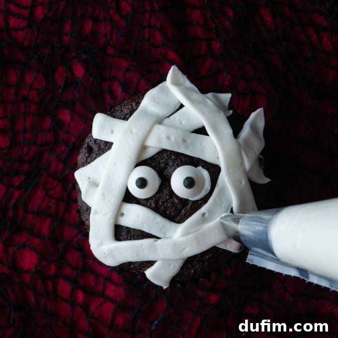 A chocolate cupcake midway through mummy decoration, showcasing the placement of candy eyes and the crisscross pattern of white frosting strips to form bandages.