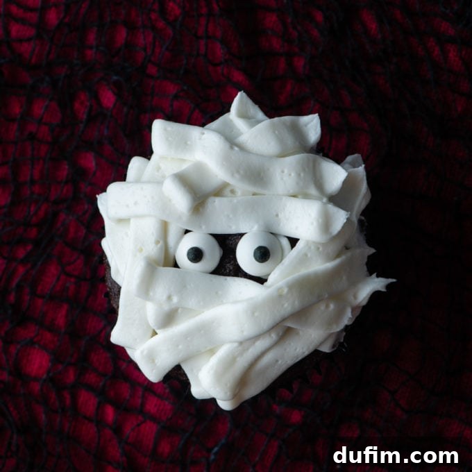 A fully decorated mummy cupcake, complete with candy eyeballs and intricate white frosting strips, resting on a red and black surface, ready to be enjoyed as a Halloween treat.