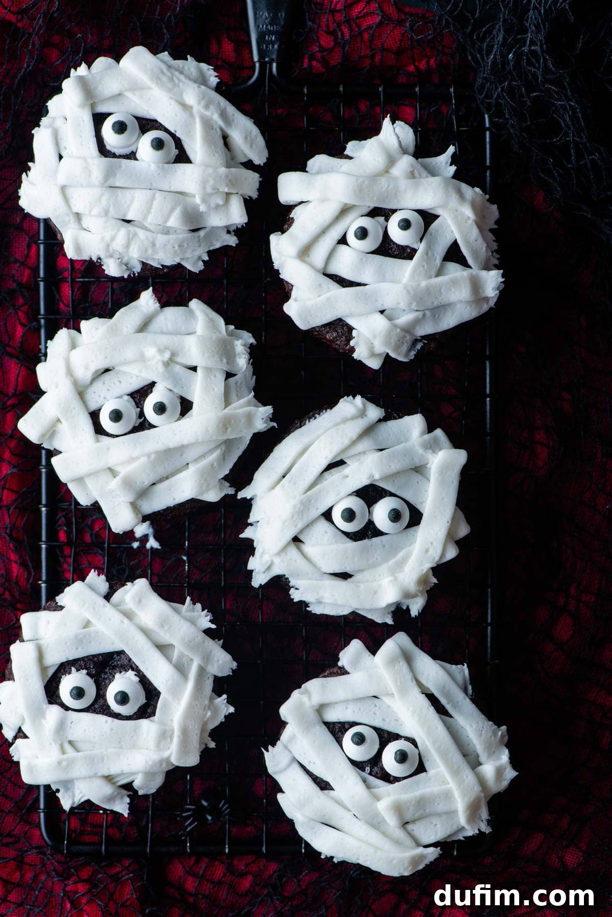 Freshly baked and decorated mummy cupcakes cooling on a wire rack above a black and red surface, showcasing their delicious appearance before serving.