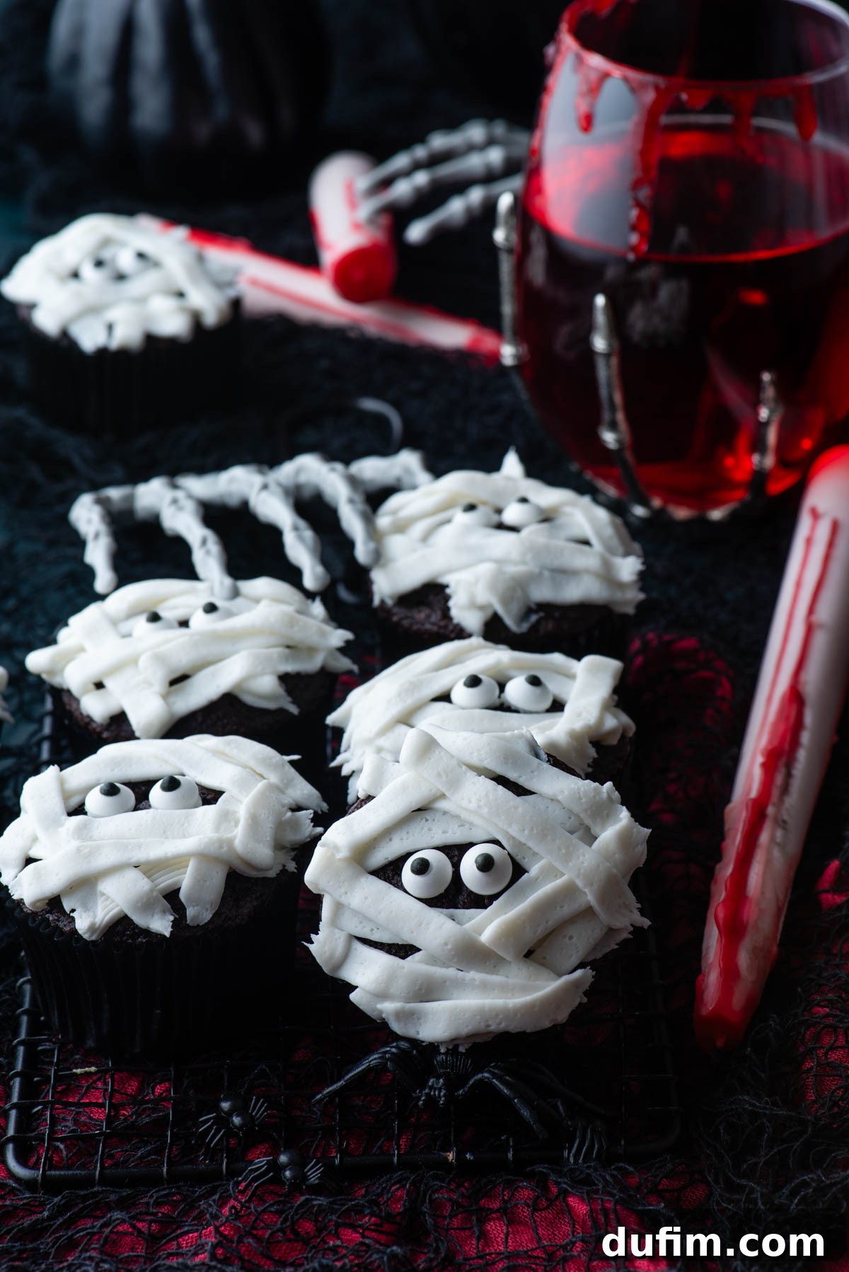 Close-up of mummy cupcakes on a black and red surface, featuring a glass of red liquid, a skeleton hand, and scattered plastic spiders, highlighting their festive Halloween presentation.