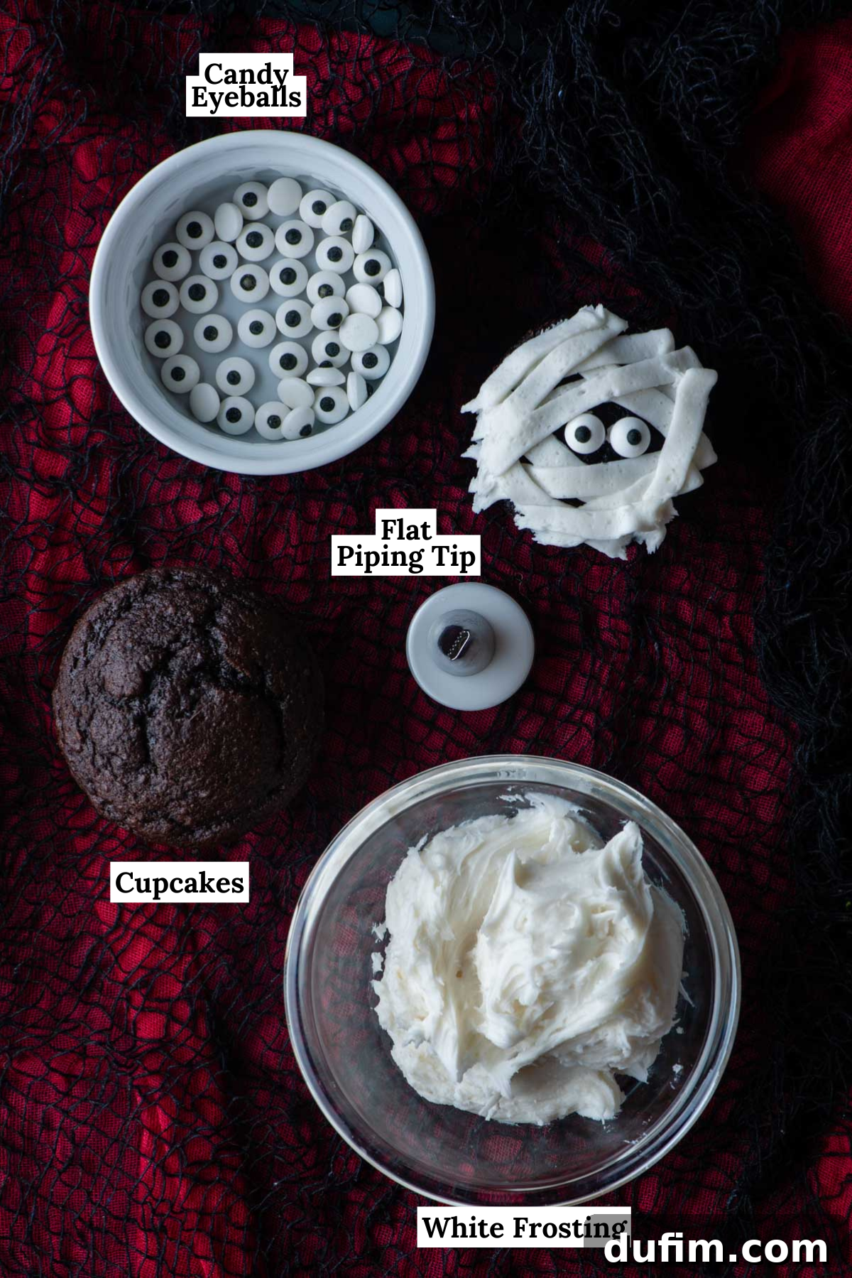 Ingredients for making mummy cupcakes laid out on a black and red surface, including baked chocolate cupcakes, a bowl of white frosting, candy eyeballs, and a flat piping tip, ready for decoration.