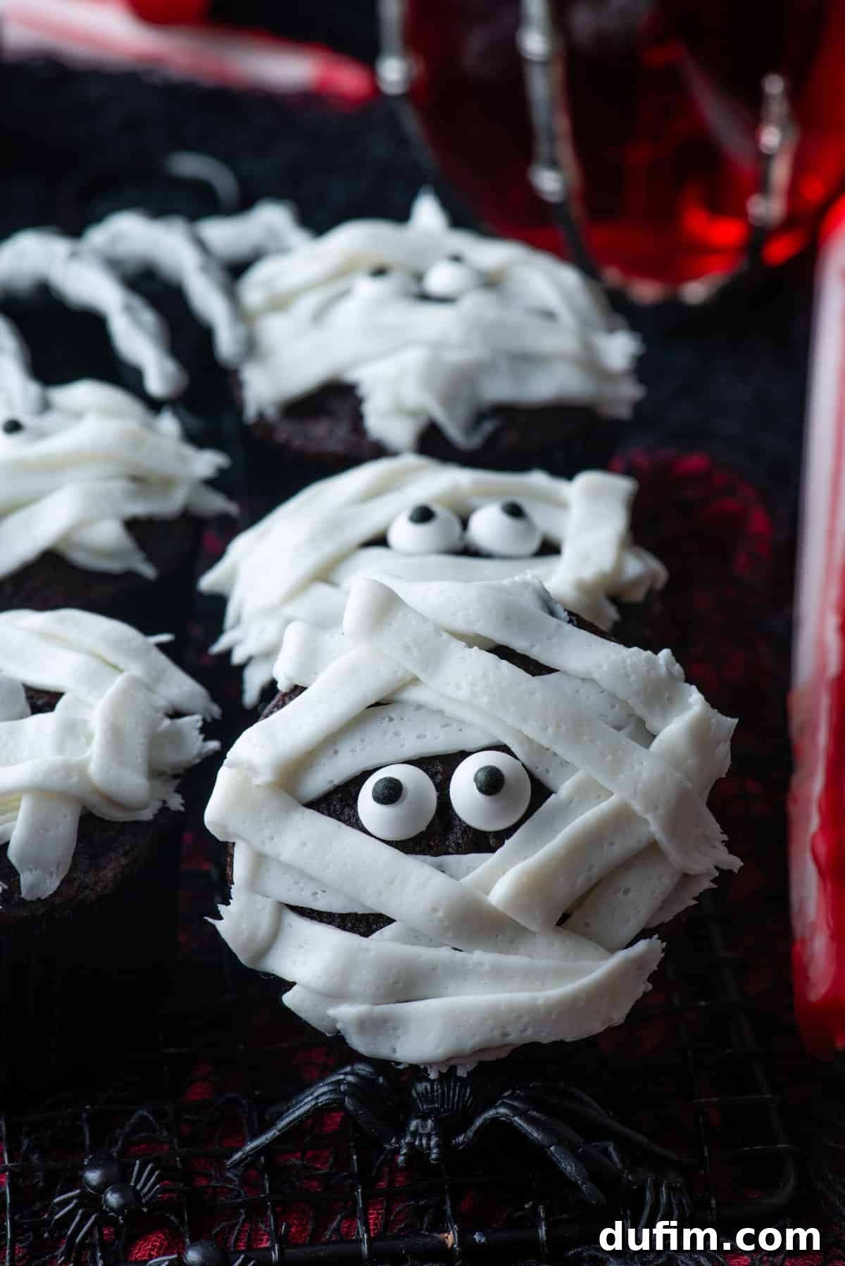 A batch of baked mummy cupcakes displayed on a black and red surface, featuring a glass of red liquid, a skeleton hand, and small black plastic spiders, setting a festive Halloween mood.