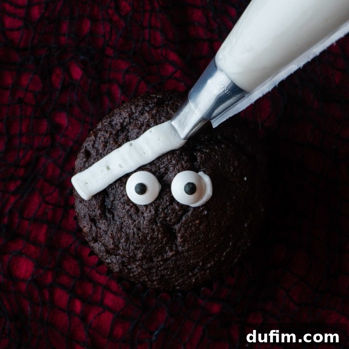 A chocolate cupcake on a red and black surface, with candy eyeballs placed in the center, and a white strip of frosting being piped diagonally across the top, beginning the mummy decoration.