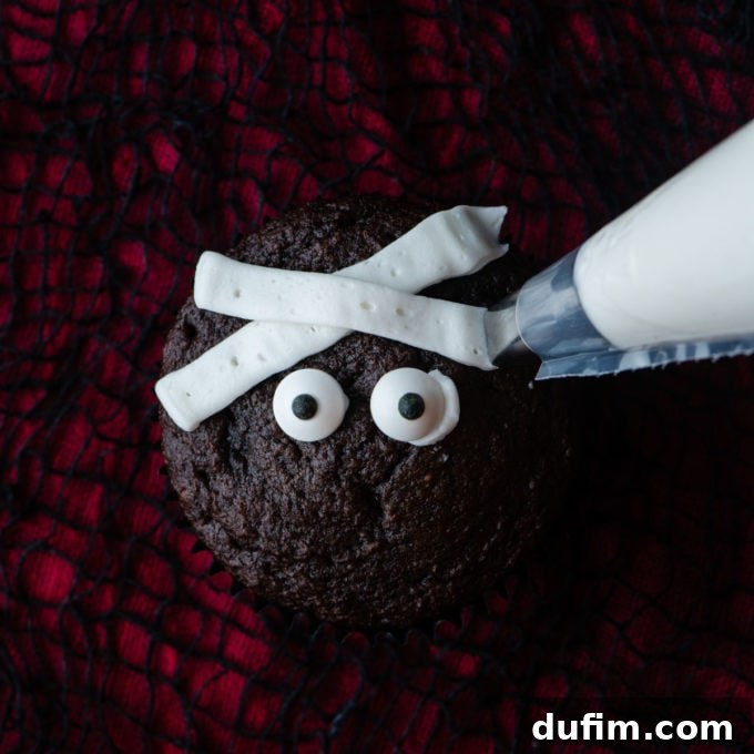 A chocolate cupcake on a red and black surface, with candy eyeballs centered and a second diagonal white frosting strip piped over the first, creating a layered mummy bandage effect.