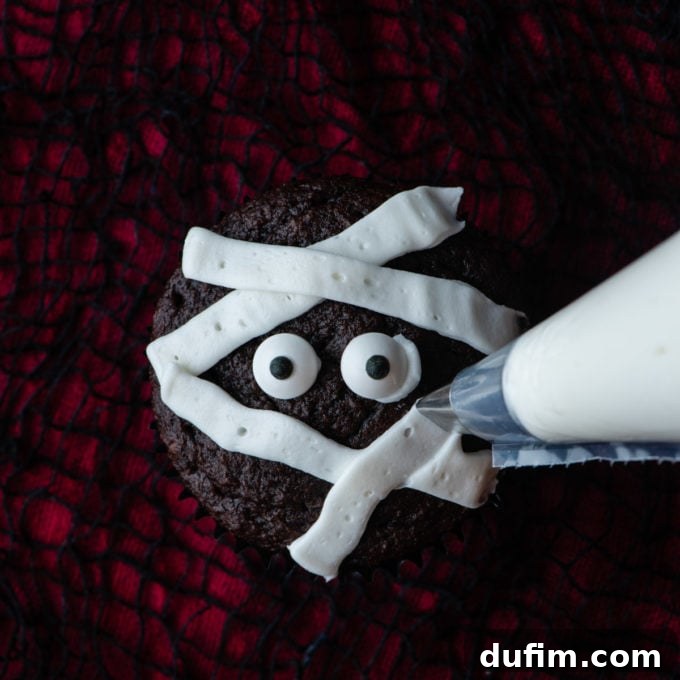 A chocolate cupcake on a red and black surface with candy eyes in the middle, showing more white frosting strips being piped crisscross to enhance the mummy appearance.