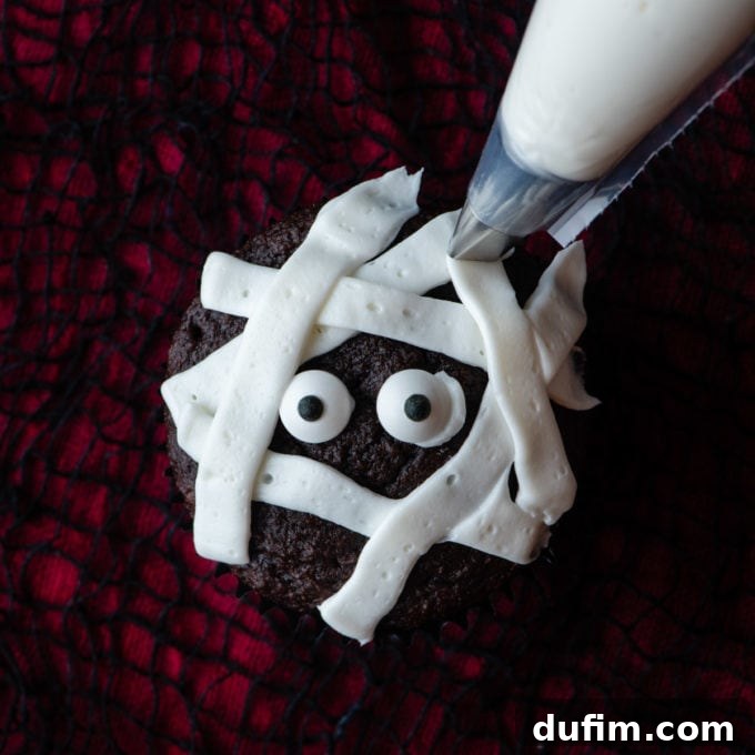A chocolate cupcake midway through mummy decoration, showcasing the placement of candy eyes and the crisscross pattern of white frosting strips to form bandages.
